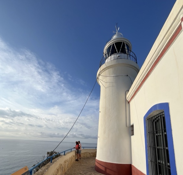 A white clifftop lighthouse with blue trim overlooking a calm sea under a clear sky, two people at the railing